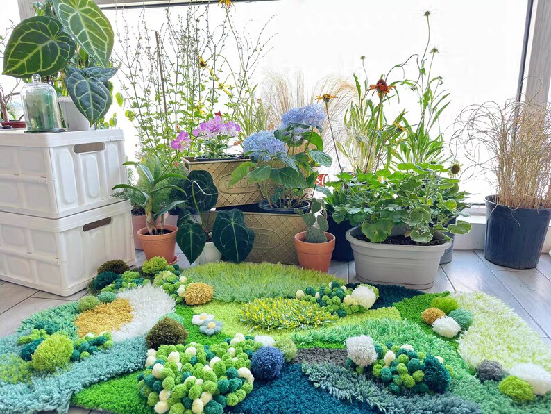 May include: A green, blue, and white rug with a textured, fluffy surface. The rug is decorated with pom-poms and resembles a garden scene. Several potted plants are visible in the background.