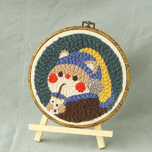 May include: A circular, embroidered artwork depicting a cartoon character wearing a blue hat and a yellow arc. The character has red cheeks and holds a small item. The artwork is set in a wooden hoop and displayed on a small wooden easel.