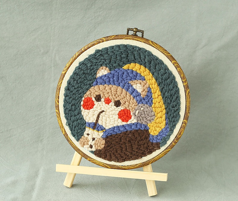 May include: A circular, embroidered artwork depicting a cartoon character wearing a blue hat and a yellow arc. The character has red cheeks and holds a small item. The artwork is set in a wooden hoop and displayed on a small wooden easel.
