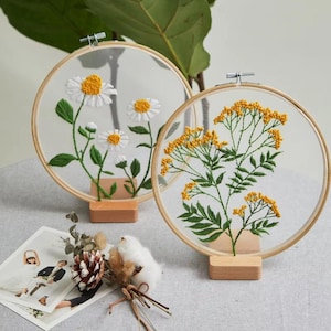 May include: Two circular embroidery hoops displaying floral designs. One shows white daisies with yellow centres and green stems. The other has yellow flowers and green leaves. Both are on wooden stands.