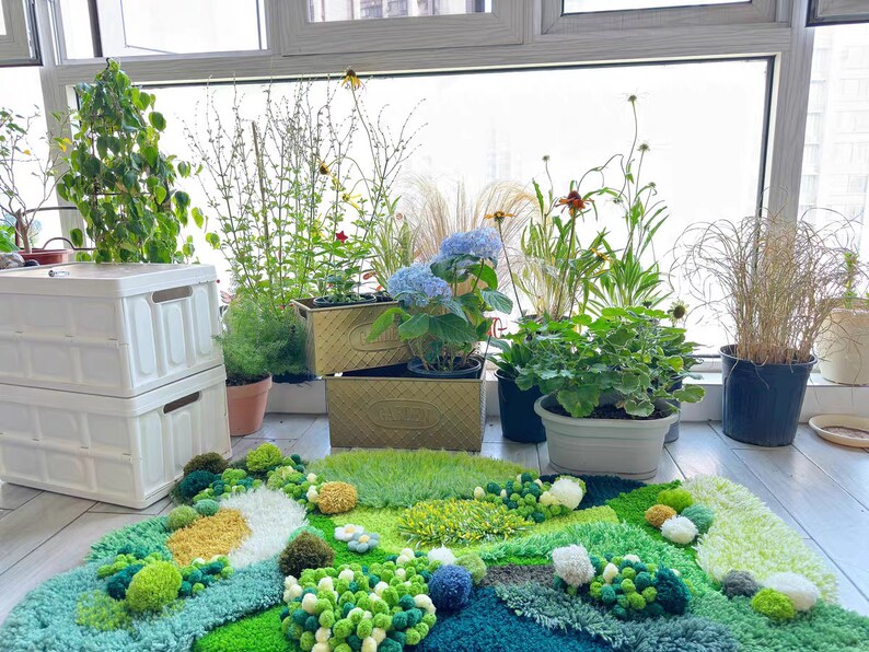 May include: A green, blue, and white rug with a variety of textures and shapes. The rug is made of yarn and has a variety of different colors and textures. The rug is in front of a window with plants in pots.