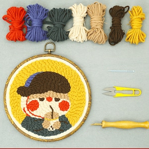 May include: A punch needle craft kit with a circular, framed artwork depicting a cartoon character wearing a hat and scarf. The kit includes colourful yarn, a needle, scissors, and a punch needle tool. The artwork is approximately 15 cm in diameter.
