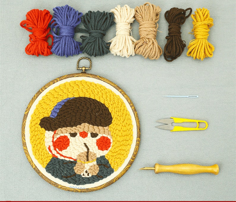 May include: A punch needle craft kit with a circular, framed artwork depicting a cartoon character wearing a hat and scarf. The kit includes colourful yarn, a needle, scissors, and a punch needle tool. The artwork is approximately 15 cm in diameter.