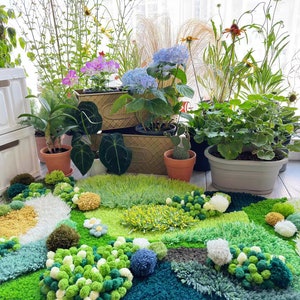 May include: A green, blue, and yellow rug with a variety of textures and shapes. The rug is made of yarn and has a fluffy, pom-pom texture. The rug is surrounded by potted plants, including a green plant with white flowers, a green plant with pink flowers, and a green plant with purple flowers.