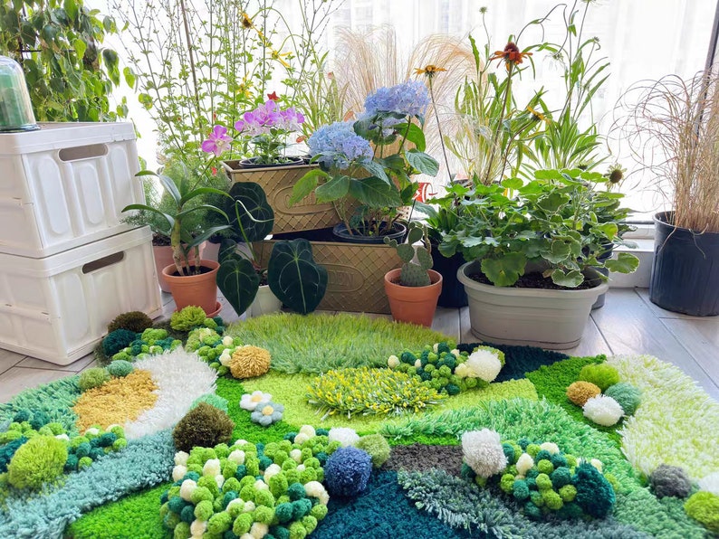 May include: A green, blue, and yellow rug with a variety of textures and shapes. The rug is made of yarn and has a fluffy, pom-pom texture. The rug is surrounded by potted plants, including a green plant with white flowers, a green plant with pink flowers, and a green plant with purple flowers.