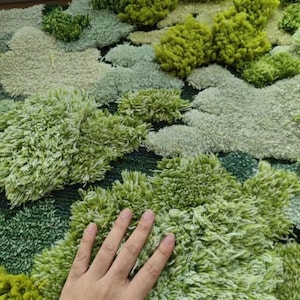 May include: A close-up of a green and white rug with a textured, fluffy surface. The rug is made of different shades of green and white, creating a realistic moss-like effect.