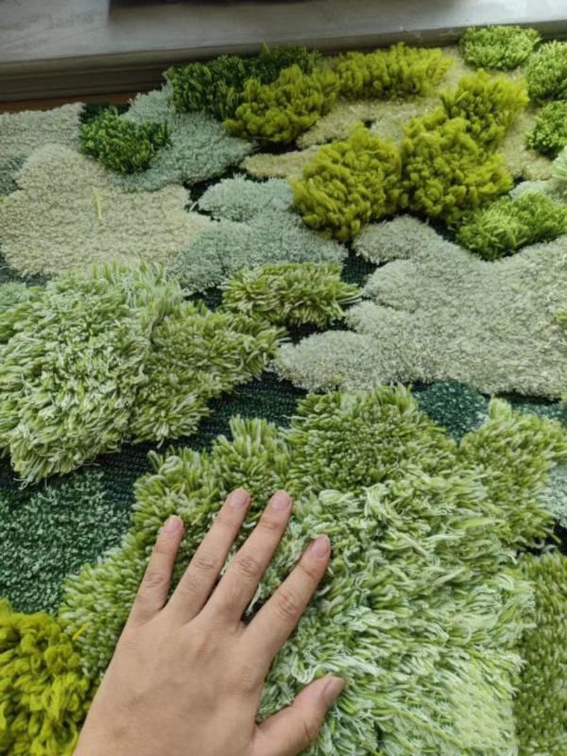 May include: A close-up of a green and white rug with a textured, fluffy surface. The rug is made of different shades of green and white, creating a realistic moss-like effect.