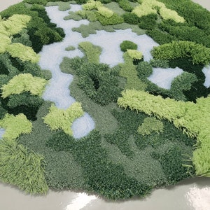 May include: A green and blue rug with a textured, fluffy surface. The rug is shaped like a circle and has a design that resembles a landscape with hills and valleys.