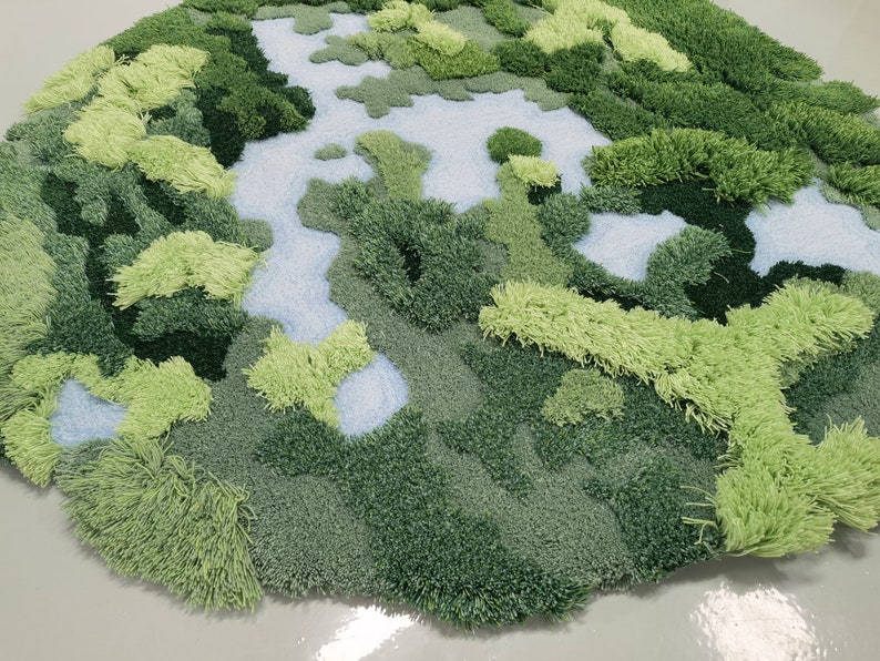 May include: A green and blue rug with a textured, fluffy surface. The rug is shaped like a circle and has a design that resembles a landscape with hills and valleys.