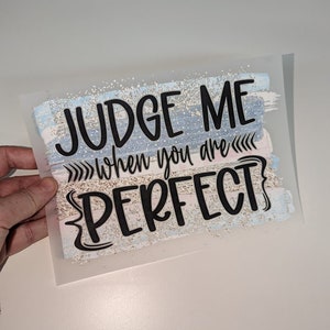 May include: A white iron-on transfer with a blue, pink and white watercolour background. The text "JUDGE ME when you are PERFECT" is in black lettering.