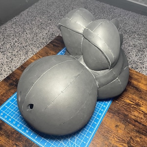 Peut inclure: Une sculpture en mousse grise en plusieurs parties, peut-être un casque ou un couvre-chef, avec des coutures visibles et une forme arrondie. L'objet est sur une surface en bois avec un tapis de coupe bleu.