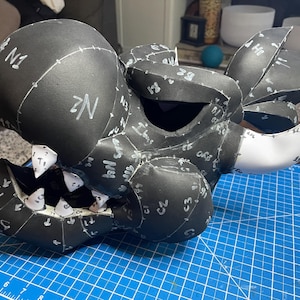 May include: A black dragon head with white markings and teeth, constructed from foam. The head is partially assembled, with visible seams and numbers. The dragon head is on a blue cutting mat.