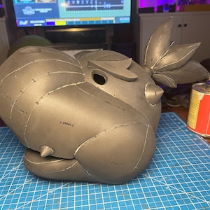 May include: A gray, unfinished cosplay helmet with a stylized design, featuring a prominent head shape, eye opening, and leaf-like accents. The helmet is marked with labels and sits on a blue grid cutting mat.