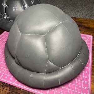 Puede incluir: Un objeto gris en forma de cúpula con paneles segmentados y un borde redondeado, que se asemeja a un caparazón de tortuga. El objeto está colocado sobre una esterilla de corte rosa con marcas de medición. Otro objeto similar es visible en el fondo.