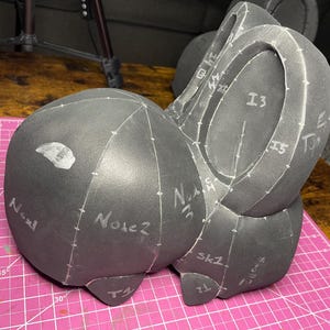 Puede incluir: Forma de cabeza de espuma gris oscuro con marcas y texto en blanco, incluyendo "Nose 2" e "I3". La forma está sobre una esterilla de corte rosa. La forma de la cabeza es probablemente para manualidades o diseño.
