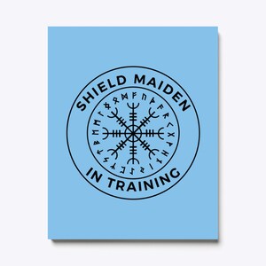 以下が含まれることがあります： 薄い青色の背景に、黒い円の中に「Shield Maiden in Training」という文字と、周囲にルーンが描かれた黒いコンパスが描かれています。