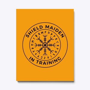 以下が含まれることがあります： 黄色の背景に、黒い円の中に「Shield Maiden in Training」という文字と、周囲にルーンが描かれた黒いコンパスが描かれています。