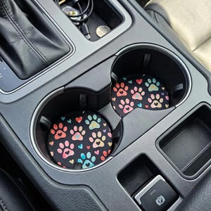 May include: Two circular car cup holder coasters featuring a black background with a repeating pattern of colorful paw prints and small hearts in shades of pink, orange, yellow, and blue, fitted inside the center console of a vehicle.