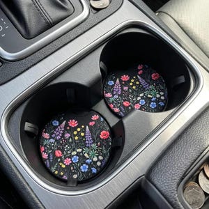 May include: Two car coasters with a black background and a colorful floral pattern. The coasters feature red poppies, purple lupine, blue wildflowers, and yellow accents. They are placed in the car's cup holders.