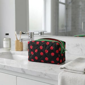 May include: Black quilted cosmetic bag with a pattern of red apples. The bag has a green zipper and trim. The bag is sitting on a white countertop in a bathroom setting.