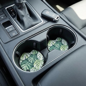 May include: Two car coasters with a tropical leaf pattern in a car's cup holders. The coasters feature a design of green and white monstera leaves. A car key and gear shift are also visible.