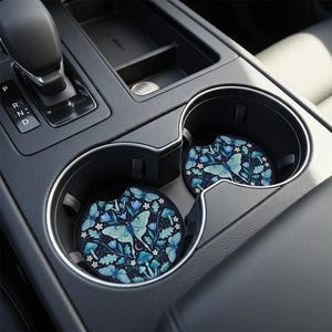 May include: Two circular car cup holder coasters featuring a dark navy blue background with a repeating pattern of light blue luna moths, small white flowers, blue mushrooms, and green oak leaves arranged in a symmetrical botanical design.