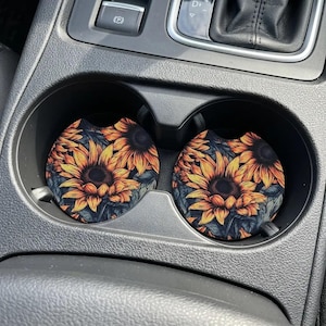 May include: Two circular car cup holder inserts featuring a vibrant sunflower pattern with bright yellow and orange petals against a dark, moody background. These round coasters are designed to fit snugly into the console of a vehicle.