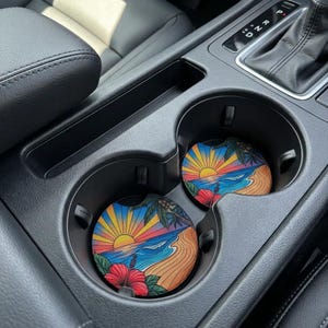 May include: Two car coasters with a tropical beach scene. The coasters feature a vibrant sunset over the ocean, palm trees, and red hibiscus flowers. They are placed in the cup holders of a car.