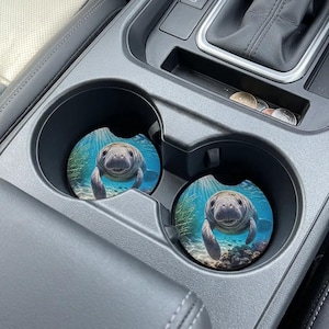May include: Two circular car cup holder inserts featuring a high-resolution photographic print of a manatee swimming underwater. The image displays the manatee in clear blue water with sunlight filtering from above, surrounded by green aquatic plants.
