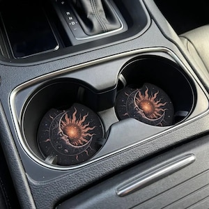 May include: Two car coasters with a celestial design featuring a sun and moon. The coasters are black with a detailed illustration in shades of orange, brown, and white. They are placed in the cup holders of a car.