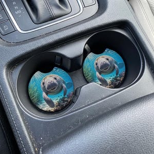 May include: Two round car coasters featuring a smiling manatee swimming underwater. The coasters have a blue background with coral and sea plants. The manatee is gray and has a friendly expression.