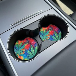 May include: Two car coasters with a colorful tie-dye design and green cannabis leaves. The coasters are placed inside the car's cup holders, with a silver trim and a black interior.