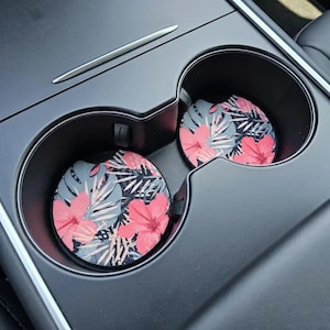 May include: Two car coasters with a tropical floral design. The coasters feature a pattern of pink hibiscus flowers, gray leaves, and black accents. They are placed inside a car's cup holders, offering a decorative and functional accessory.