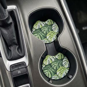 May include: Two circular car cup holder inserts featuring a repeating pattern of green and white monstera leaves. The coasters are positioned inside the black plastic cup holders of a vehicle center console, adjacent to the gear shift and buttons.