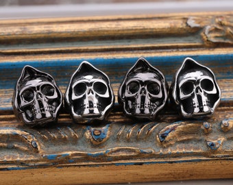 May include: Four silver skull-shaped charms with a hooded design. The charms are arranged in a row on a wooden surface.