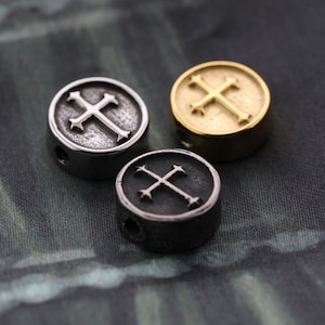 Könnte beinhalten: Drei runde Metallperlen mit Kreuzdesign. Die Perlen sind silber, gold und schwarz.