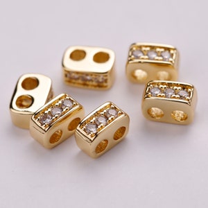 May include: Gold-colored rectangular jewelry beads with two holes. Some beads have a row of small, clear crystals. The beads are arranged on a white surface, showcasing their shiny, reflective surfaces. These beads are ideal for jewelry making.