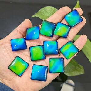 May include: A collection of 10 iridescent square-shaped buttons with a blue and green color shift. The buttons are arranged in a semi-circle on a hand.