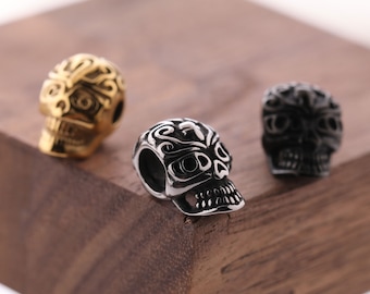 Op de afbeelding: Drie schedelvormige kralen met ingewikkelde ontwerpen. De kralen zijn gemaakt van metaal en zijn goud, zilver en zwart van kleur.