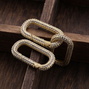 May include: Two gold-coloured, rectangular clasps with a pave setting of small, clear gemstones. The clasps are interlocked, showcasing their design. The background is a dark, wooden surface.