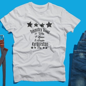 May include: White t-shirt with black text that reads "Dmitry Bivol" and his stats: 21 Wins, 0 Losses, 0 Draws, and Kyrgyzstan. The shirt is paired with blue jeans, a brown belt, and a smartphone with earbuds.