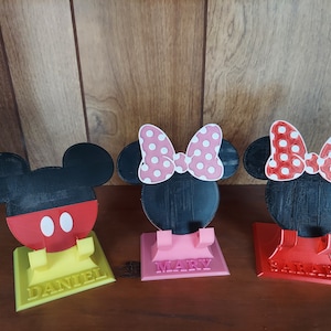 May include: Three 3D printed phone stands featuring Mickey Mouse and Minnie Mouse. The stands are yellow, pink, and red. The stands have the names "Daniel", "Mary", and "Sarah" printed on them.