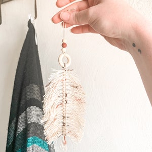 K&ouml;nnte beinhalten: Eine wei&szlig;e Makramee-Feder mit braunen Perlen und einem Holzring, der an einer Schnur h&auml;ngt.