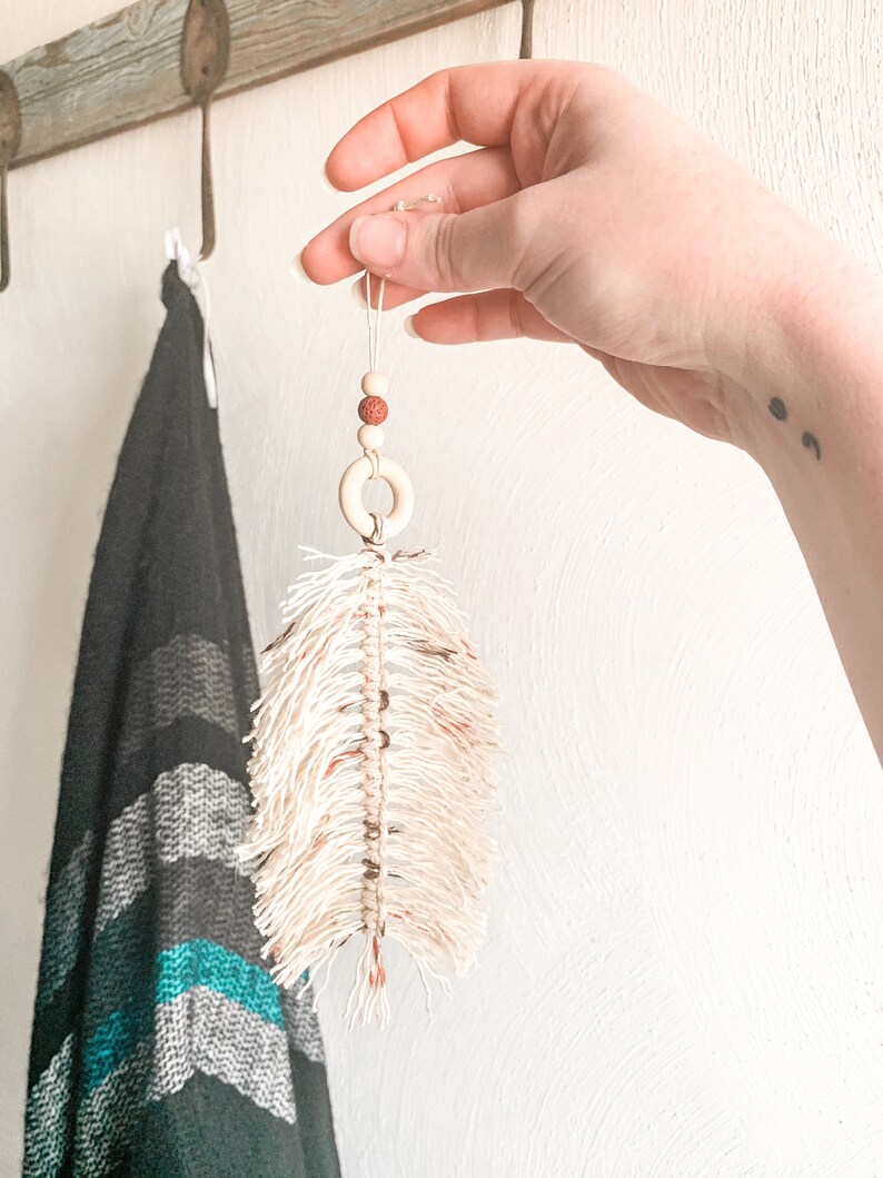 K&ouml;nnte beinhalten: Eine wei&szlig;e Makramee-Feder mit braunen Perlen und einem Holzring, der an einer Schnur h&auml;ngt.