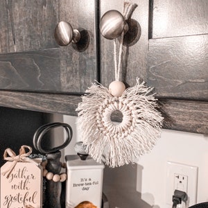 May include: A white macrame wreath with fringe hanging from a cabinet door knob. The wreath is made of natural fibers and has a rustic, farmhouse style. The cabinet door is dark brown wood with silver knobs. There is a sign that says "gather with grateful hearts" and a small white ceramic jar that says "It's a Brew-tea-ful day" on a shelf below the wreath.