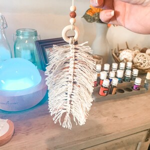 Op de afbeelding: Een witte macramé veren autoluchtverfrisser met een houten ring en bruine kralen die aan een touwtje hangen.