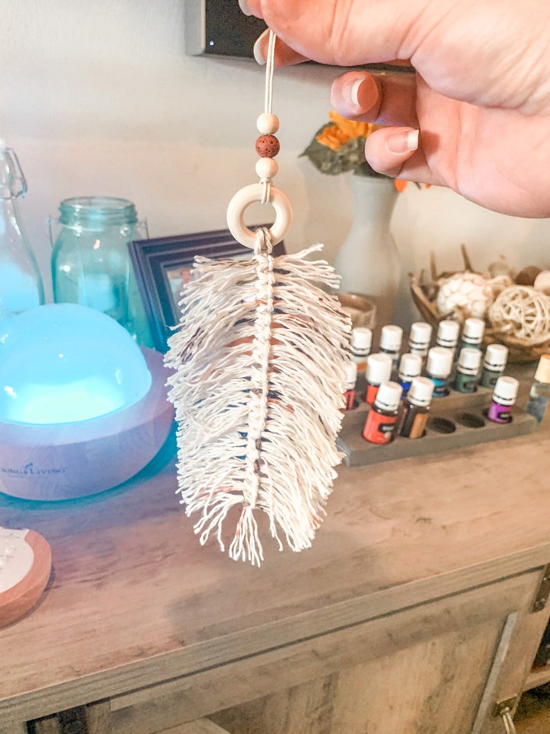 K&ouml;nnte beinhalten: Ein wei&szlig;er Makramee-Feder-Lufterfrischer f&uuml;r das Auto mit einem Holzring und braunen Perlen, die an einer Schnur h&auml;ngen.