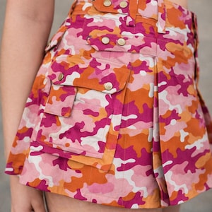 May include: A pink, orange, and white camouflage print skirt with multiple pockets and gold-colored button details. The skirt has a pleated design and a high waist. The overall theme is fashion and clothing.