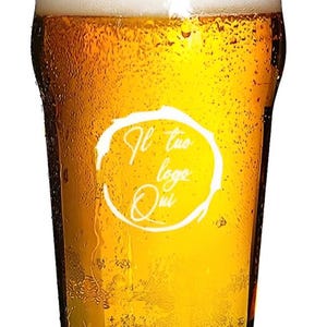 Può includere: Un bicchiere di birra con una schiuma bianca in un bicchiere di pinta trasparente. Il bicchiere ha un cerchio bianco con il testo "Il tuo logo qui" sul lato.