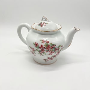 May include: A white porcelain teapot featuring a delicate floral pattern of small red berries and green leaves. The teapot has a curved handle, a short spout, and a matching lid with a gold-trimmed scalloped edge and a small finial handle.